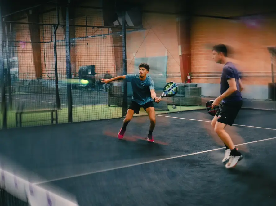 A dynamic, motion-blur action shot of two men playing padel on a black indoor court. The player in the background, wearing a blue shirt, stretches out to hit a neon green ball with his racket, while the foreground player in a purple shirt watches the play intently.