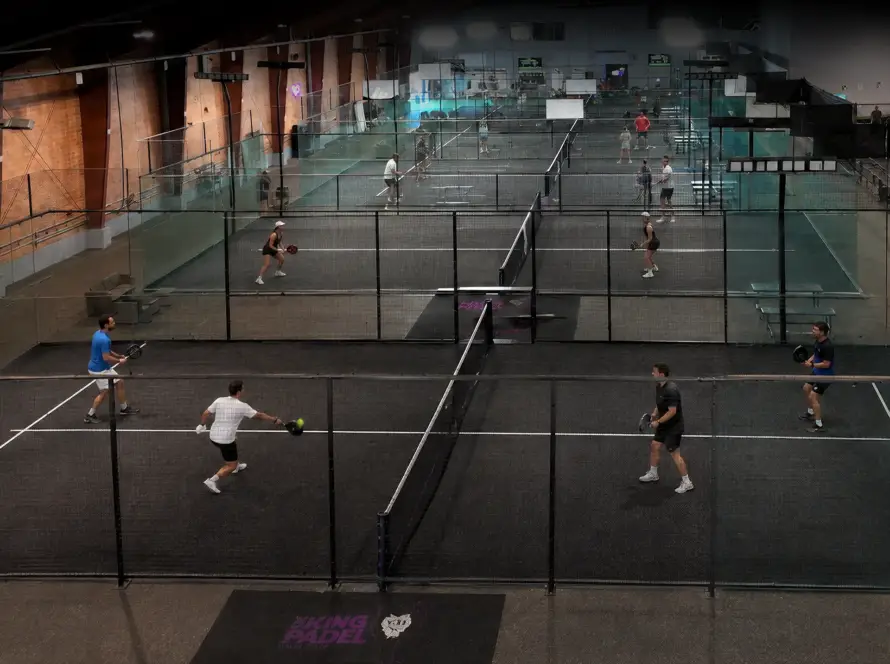 An elevated, wide-angle panoramic view of an indoor sports complex featuring multiple black padel courts lined up in a row. Several pairs in athletic wear are seen playing simultaneously on courts separated by glass walls and metal netting, with a "King's Padel" logo visible on the floor mats.