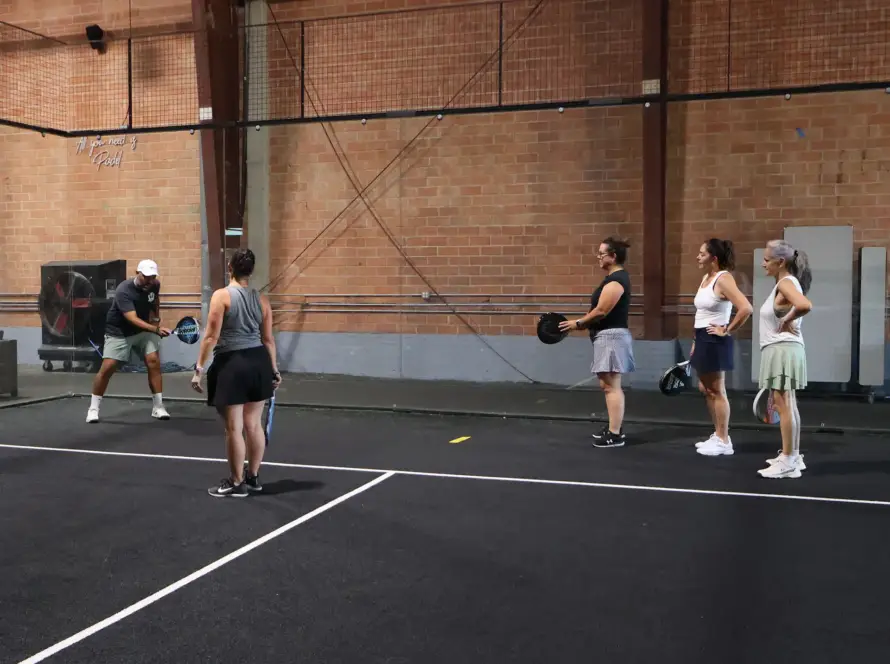 An instructor wearing a white cap and black t-shirt gives tactical directions on a padel court to a lineup of four women in athletic wear who are listening attentively while holding their rackets. A high exposed-brick wall stands in the background.