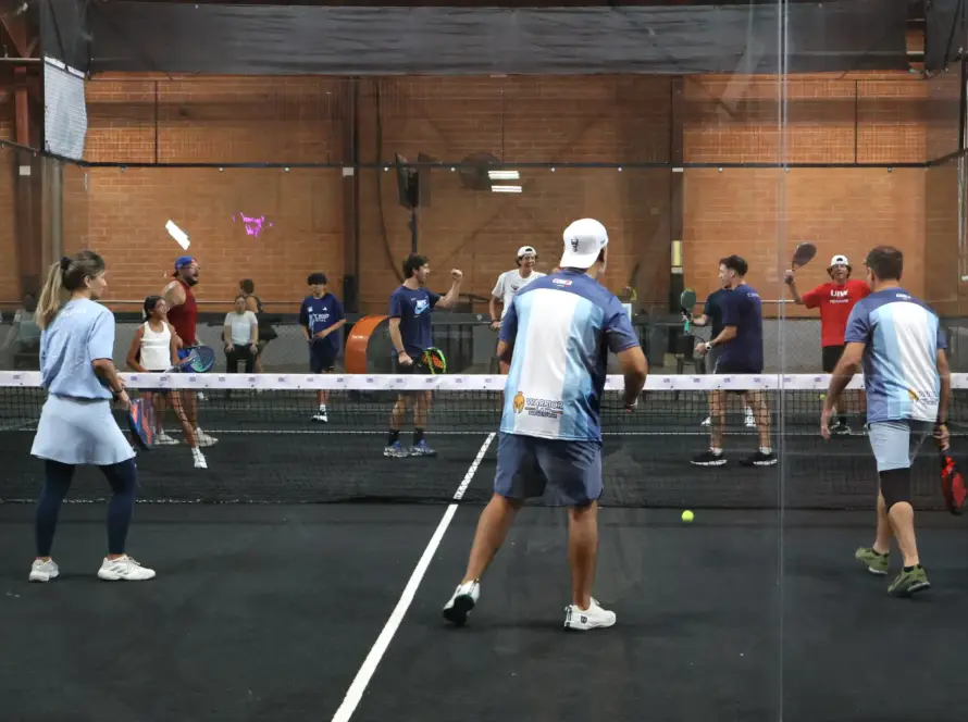A behind-the-glass view of multiple groups of players chatting and high-fiving over the nets during an indoor padel tournament. The shot captures a lively, social club environment with exposed brick walls in the background.