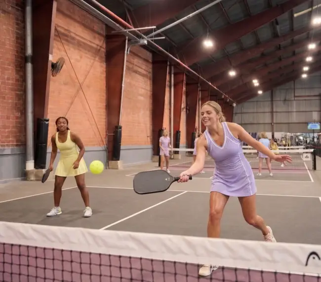 Women playing pickleball, laughing, energetic. Pink gear if available from past events. 